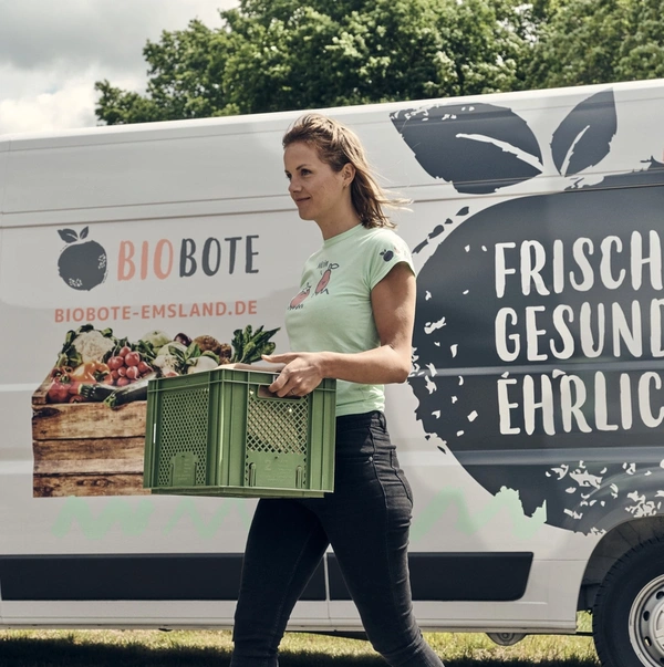 KI generiert: Eine Frau trägt eine grüne Kiste vor einem Lieferwagen mit der Aufschrift "BIOBOTE" und "FRISCH, GESUND, EHRLICH".