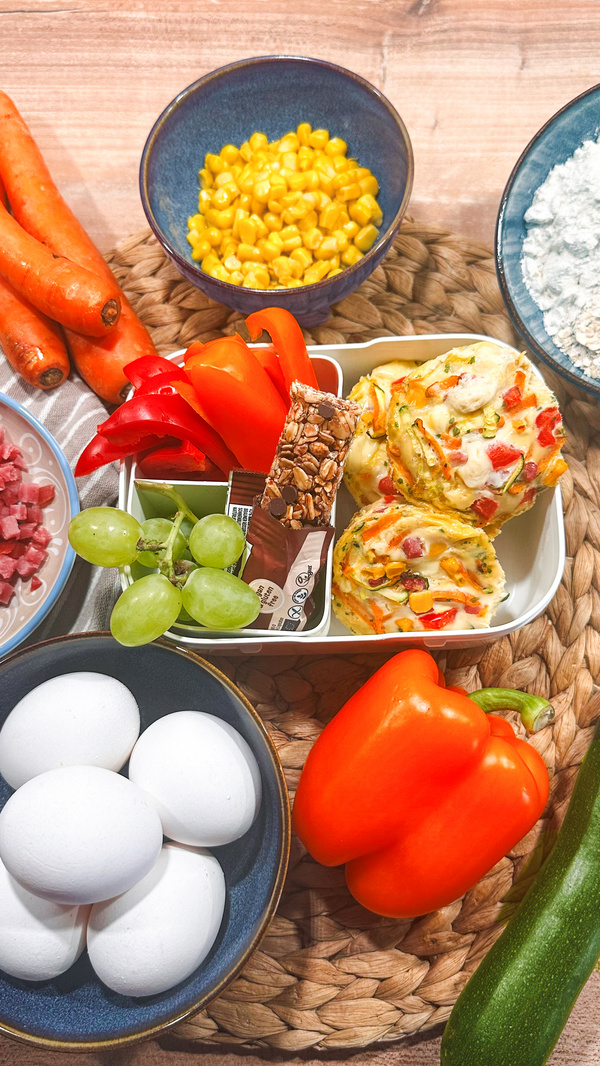 KI generiert: Frische Zutaten und Gemüse, darunter Paprika, Eier, Möhren, Mais und Muffins, arrangiert auf einem Tisch.