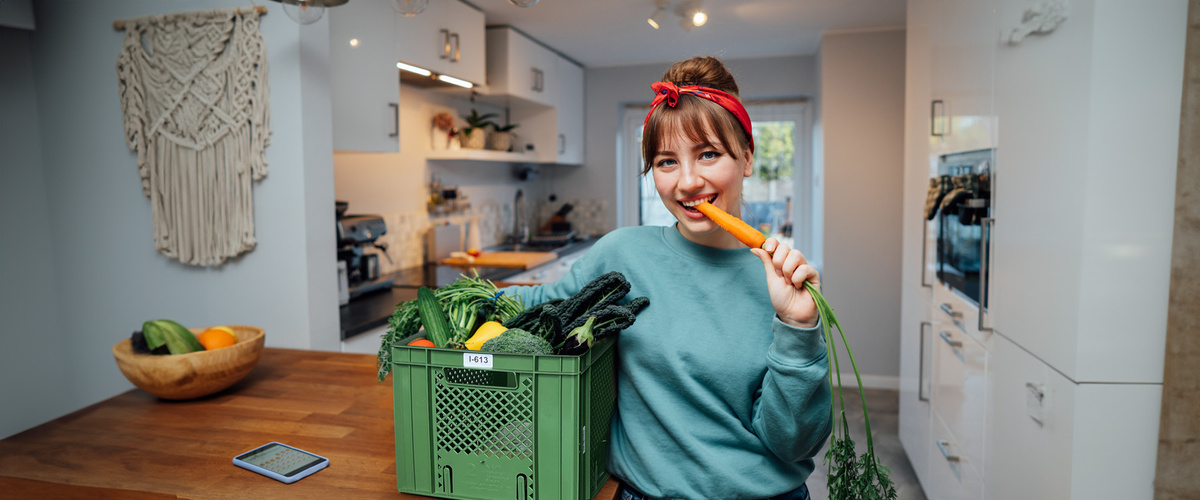 KI generiert: Eine Frau in der Küche beißt lächelnd in eine Karotte neben einer Kiste mit Gemüse.