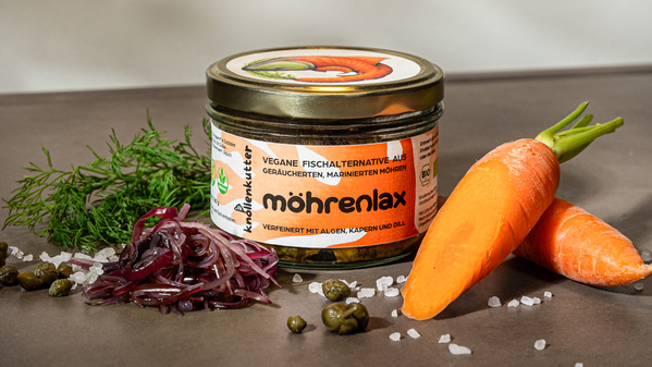 KI generiert: Ein Glas "möhrenlax" mit Karotten, Dill, Zwiebeln und Kapern auf einem Tisch. Text: "Möhrenlax".