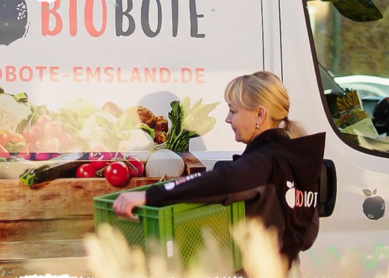 KI generiert: Eine Frau liefert eine grüne Kiste; im Hintergrund ein Biobote-Lieferwagen. Text: "BIOBOTE", "BIOBOTE-EMSLAND.DE".