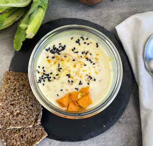 KI generiert: Das Bild zeigt ein Glas mit cremiger Suppe, garniert mit schwarzen Sesamsamen und orangen Stücken, daneben liegen Brotscheiben und verschiedene frische Gemüsearten wie Blumenkohl und Süßkartoffeln. Es vermittelt einen rustikalen, gesunden Eindruck.