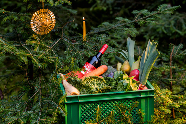 KI generiert: Grüne Kiste mit Gemüse und einer Weinflasche vor einem Weihnachtsbaum.