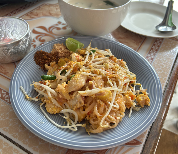 KI generiert: Ein Teller mit Pad Thai, dazu Limette, Erdnüsse, Chili und eine Beilage in einer weißen Schüssel.