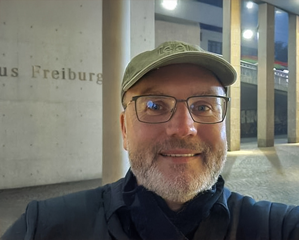 KI generiert: Ein Mann mit Brille und Mütze lächelt. Hintergrund: Säulen und Text "Freiburg" erkennbar.