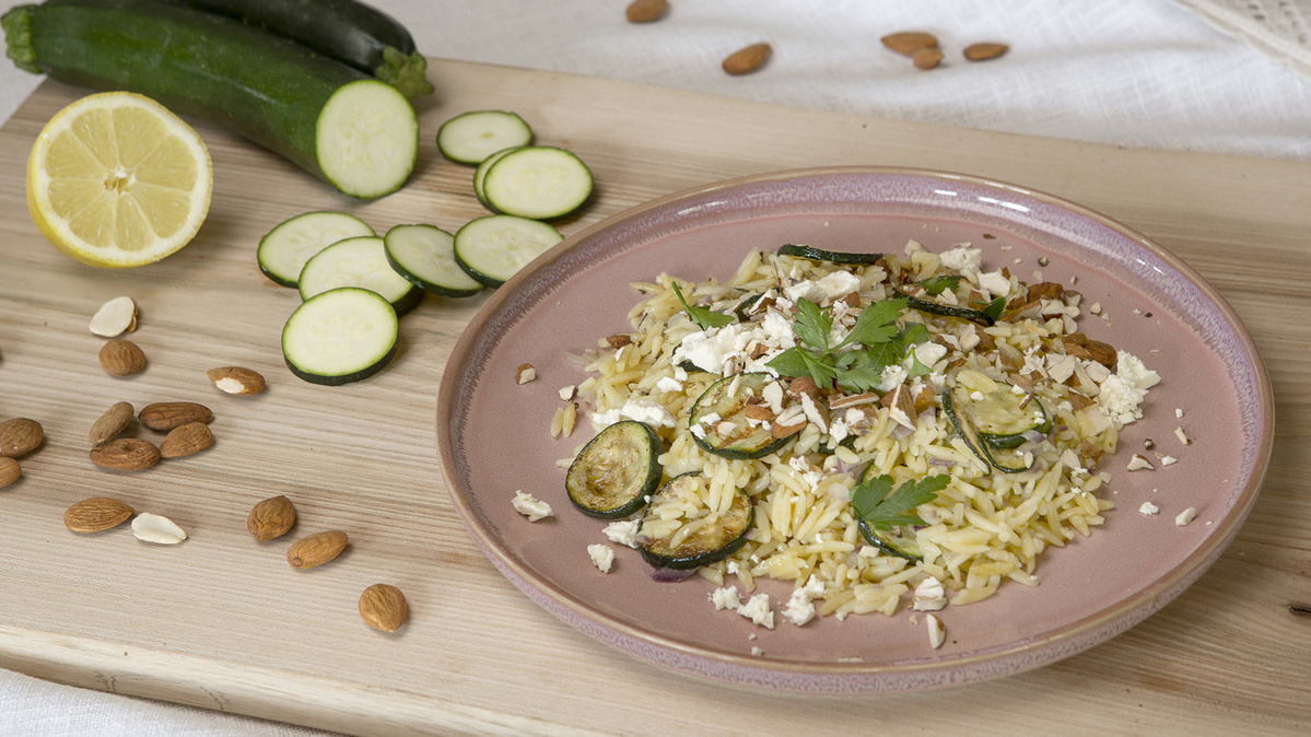 Ein Teller mit Orzo, Zucchini, Mandeln, Petersilie und Feta. Daneben Zucchinischeiben, Zitrone und Mandeln.