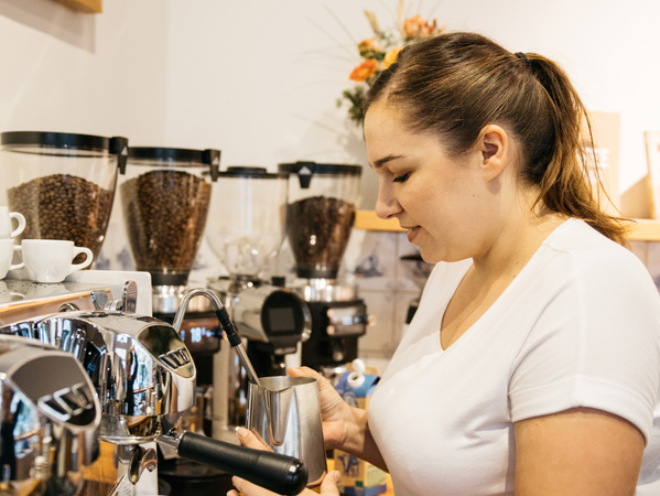 KI generiert: Das Bild zeigt ein Glas mit einer verzierten Kaffeespezialität auf einem Teller neben einer Packung Kaffee von der "Kaffeerösterei Baum". Im Hintergrund sind Kaffeebohnen zu sehen.