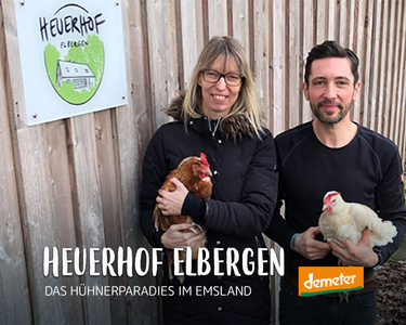 Zwei Personen halten Hühner vor einem Schild mit der Aufschrift "Heuerhof Elbergen". Text: "Das Hühnerparadies im Emsland".
