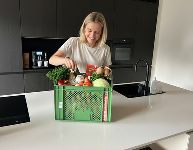 Eine junge Frau freut sich über eine Kiste mit Bio-Gemüse in einer modernen Küche. Text: "biobote.de".