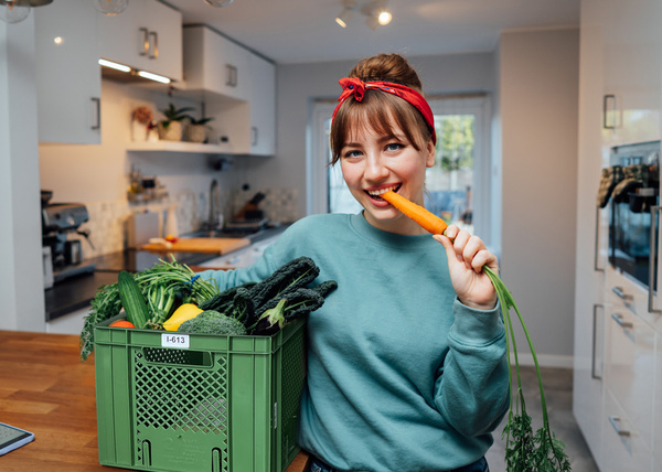 KI generiert: Eine Frau in einer Küche isst eine Karotte aus einer Gemüsebox.