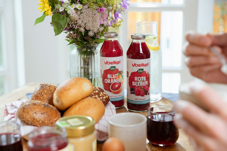 Voelkel 0,5-l-Glasmehrwegflasche, Frühstückstisch mit Bio-Getränken, Brötchen und Blumenstrauß. Text: "Voelkel bio Blut Orange", "Voelkel bio Rote Beeren".