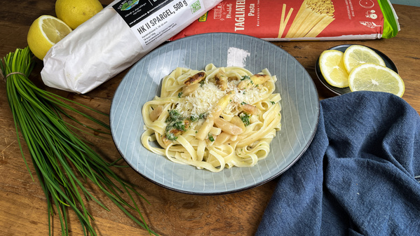 KI generiert: Ein Teller Pasta mit Spargel und Parmesan, umgeben von frischen Zutaten und Verpackungen. Text: "TAGLIATELLE".
