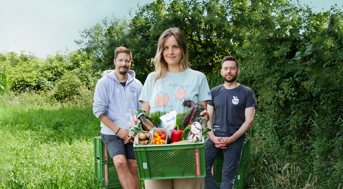 KI generiert: Drei Personen und Gemüsekiste auf Wiese, Imkerei-Logo auf T-Shirts, viel grüner Hintergrund.