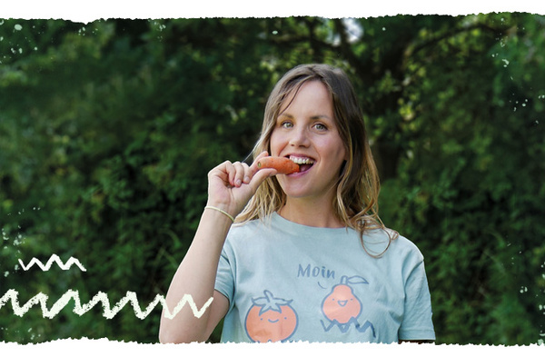 KI generiert: Eine Frau steht draußen vor einem Hintergrund mit Bäumen und beißt in eine Karotte. Sie trägt ein hellblaues T-Shirt mit einem Druck von zwei Orangen und dem Wort "Moin".