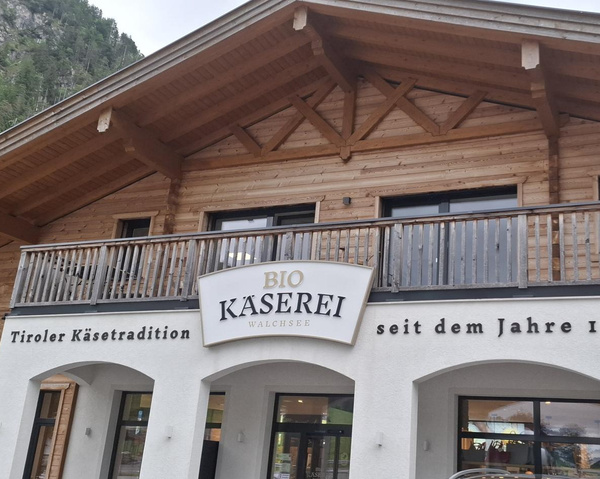 KI generiert: Das Bild zeigt die Fassade eines Bio-Käserei Ladens mit Holzverkleidung und Schildern. Text: "BIO KÄSEREI WALCHSEE Tiroler Käsetradition seit dem Jahre I".