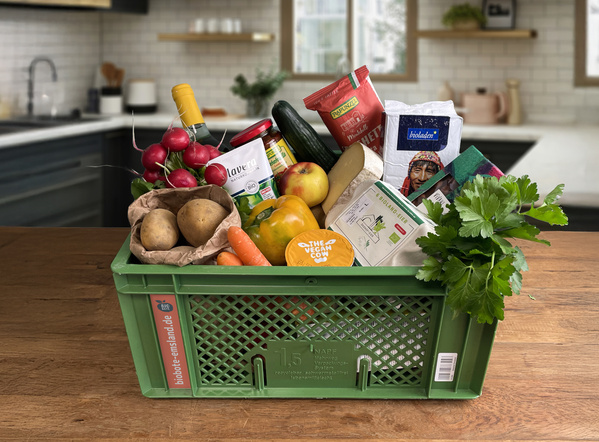 Eine gepackte grüne Bio-Kiste mit frischem Obst, Gemüse und verschiedenen Produkten auf einem Küchentisch.