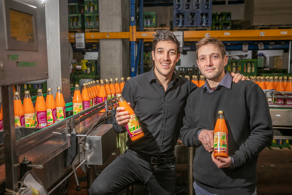 Zwei Männer in einer Fabrik halten Flaschen mit orangem Getränk. Im Hintergrund sind viele Flaschen zu sehen.